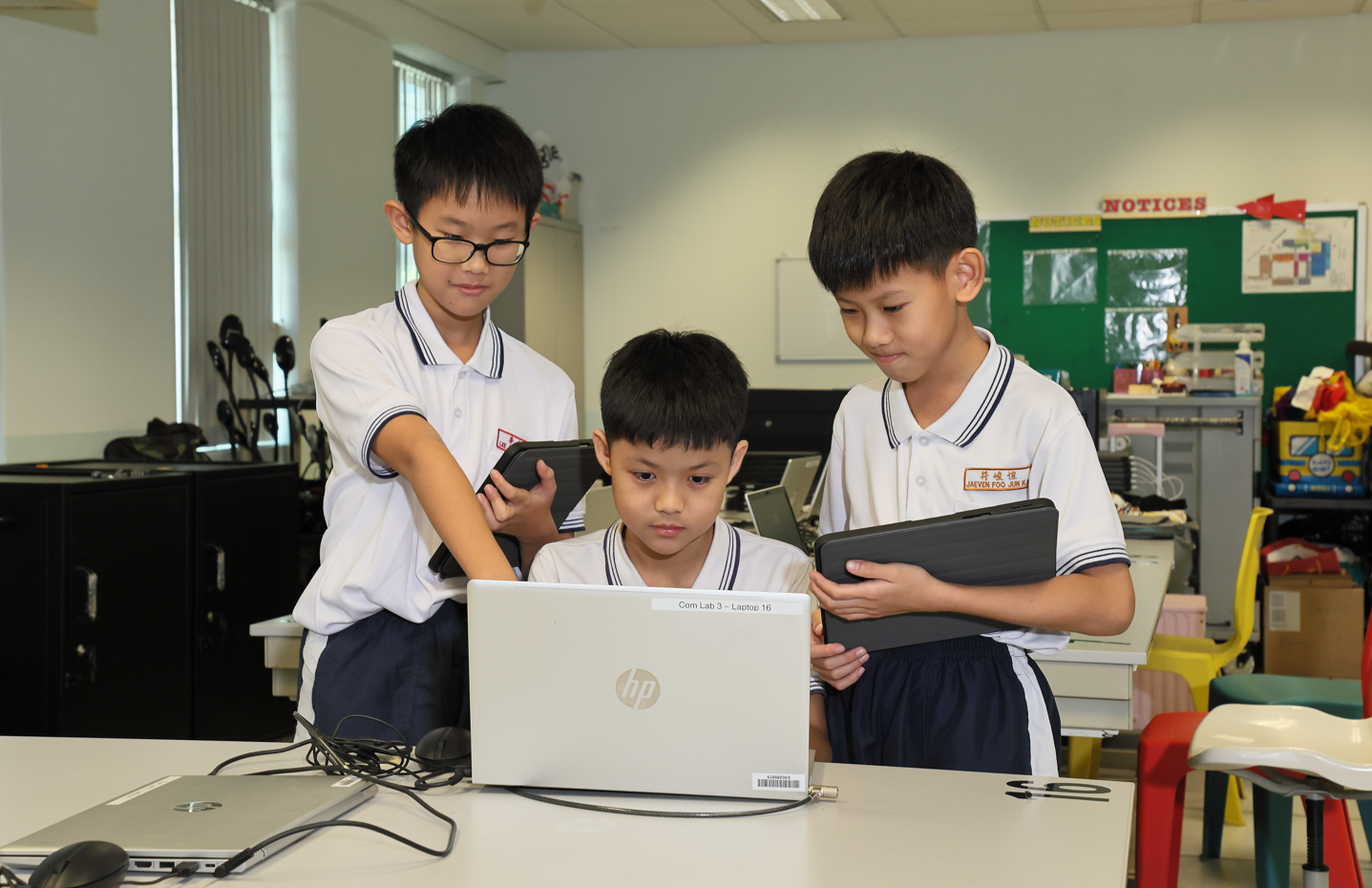 Two young boys sit at a computer desk, one wearing headphones, both looking at a monitor in the computer lab.
